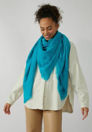 Écharpe turquoise en tissu doux, avec un design à motifs texturés et des bords à franges, enroulée lâchement autour du cou sur une chemise légère.