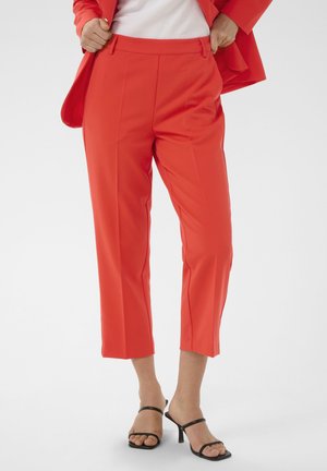 Kobieta ubrana w czerwone, krótkie, dopasowane spodnie, do kompletu marynarkę, biały top i czarne sandały na obcasie, stoi na białym tle.