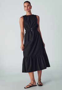 Black sleeveless dress with a gathered neckline, cinched waist featuring a silver buckle, and a tiered ruffled hem. Flat black sandals.