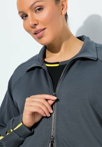 Mujer con chaqueta gris oscuro y una camiseta negra y amarilla, cerrándose la chaqueta frente a un fondo liso y claro.