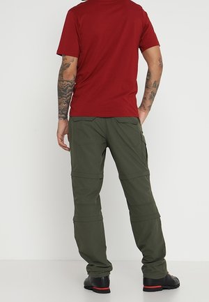 Homme portant une chemise rouge à manches courtes et un pantalon cargo vert olive avec des bottes noires, debout de dos devant un fond uni.