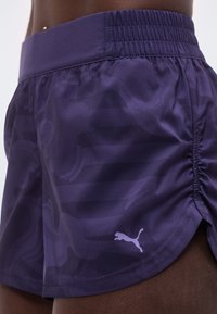 Σκούρο μωβ αθλητικό σορτς με διακριτικό μοτίβο και λογότυπο Puma, φορεμένο από άτομο με σκούρο δέρμα, που δείχνει ελαστική μέση και πτυχώσεις στο πλάι.