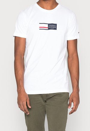 Man draagt een wit T-shirt met korte mouwen met een Tommy Hilfiger-logo op de borst en olijfgroene broek tegen een effen achtergrond.