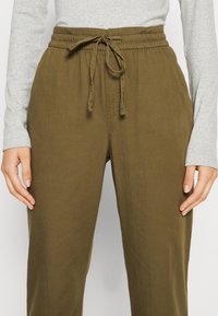 Pantalons de jogging vert olive avec une taille élastique et un lien ajustable. Le tissu semble doux et texturé, conçu pour le confort et l'ajustement.