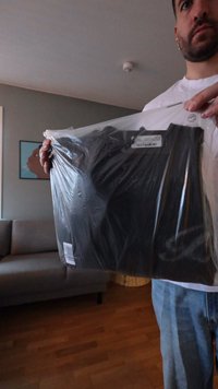 Melnas apģērba prece plastmasas iepakojumā ar svītrkoda etiķeti, ko tur persona uz neitrālas fona un gaiša koka grīdas fona.