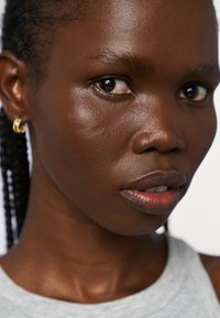 Gros plan sur un visage avec une peau lisse et foncée, présentant des cheveux tressés et de petites boucles d'oreilles en or. Porte un haut sans manches gris clair.