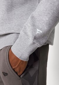 Felpa grigia con polsini a costine e un piccolo logo bianco sulla manica. Pantaloni grigi scuri con un motivo testurizzato vicino alla tasca.