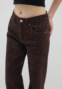 I jeans a stampa leopardata marrone presentano una vita alta, cinque tasche e una chiusura con bottone, realizzati in tessuto testurizzato con una leggera lucentezza.
