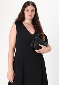 Černé bezramínkové šaty s výstřihem do V, hladká textura. Drží malou černou koženou kabelku s obloukovou rukojetí.