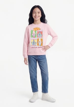 Usmívající se dívka s dlouhými černými vlasy stojí s rukou na boku, oblečená v růžové mikině s barevnými motivy oken, modrých džínách a bílých teniskách.