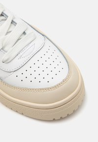 Baskets en cuir blanc avec des accents beige, semelle en caoutchouc texturé, boîte à orteils perforée et lacets plats. Présente des détails brodés près du bord.