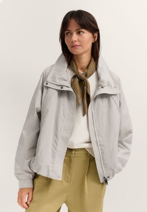 Frau, die eine hellgraue Jacke mit Reißverschluss, ein weißes Top, einen olivgrünen Schal, am Hals gebunden, und senfgelbe Hosen trägt, steht vor einem einfarbigen Hintergrund.