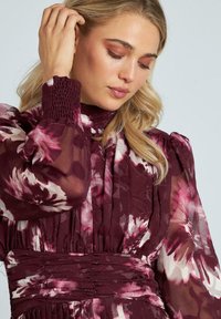 Vestido floral burdeos con mangas transparentes y abullonadas, detalle de muño en las muñecas y cintura fruncida, que muestra un patrón floral texturizado.