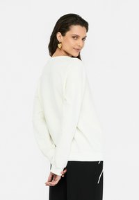 Kreemjas sweatshirt, millel on pikad varrukad, soonikkoes käised ja ümmargune kaelus, millele on sobitatud mustad püksid. Sile tekstuur ja vabalt langev lõige.