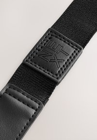 Detalizēts melnas austas auduma jostas attēls ar iešūtu melnas ādas uzliku, uz kuras ir iespiests "NEXT" logotips.