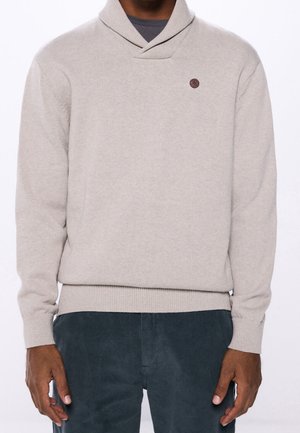 Man draagt een lichtbeige gebreide trui met sjaalkraag en een klein cirkelvormig logo op de borst, gecombineerd met donkerkleurige corduroy broek.