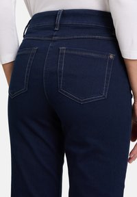 Jean en denim bleu foncé avec une taille haute, comportant deux poches arrière, des coutures contrastantes et une texture lisse.