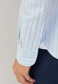 Lichtblauwe langwerpige shirt met witte en groene verticale strepen. Heeft knoopmanchetten en een gladde katoentextuur.