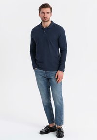 Man som står med händerna i fickorna, iklädd en marinblå långärmad polo, blå jeans och svarta läderskor mot en vit bakgrund.