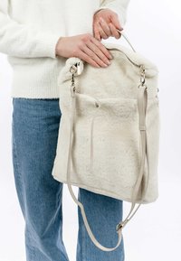 Zaino in pelliccia artificiale bianca con spallacci beige e dettagli in metallo argentato, dotato di una cerniera superiore e una tasca esterna con chiusura a coulisse.