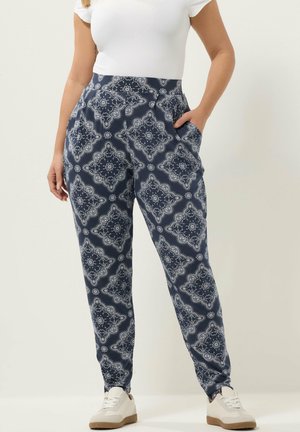 Vrouw draagt marineblauwe broek met bandana-print, wit aansluitend kort mouwshirt en witte sneakers, staat met één hand in haar zak.