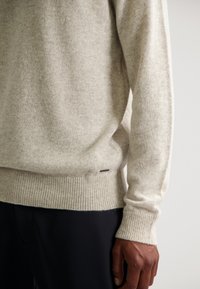 Beige gebreide trui met geribbelde zoom en manchetten, zachte textuur en subtiel patroon. Etiket zichtbaar nabij de onderste naad. Close-up van de details van het kledingstuk.