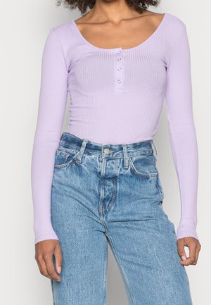 Femme portant un haut à manches longues côtelé lavande avec des boutons et un jean en denim bleu clair taille haute, debout devant un fond uni.