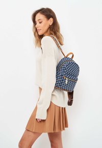 Marinblå ryggsäck med geometriskt mönster, läderdetaljer och guldfärgade detaljer. Har en böjd form och justerbara remmar.