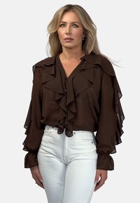 Brązowa, falbaniasta bluzka z długimi rękawami i dekoltem w kształcie V; przezroczysty materiał; w połączeniu z białymi dopasowanymi dżinsami. Subtelne guziki z przodu i przy mankietach.