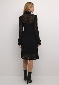 Rochie neagră din dantelă cu mâneci lungi, guler înalt și talie strânsă. Are o textură semi-transparentă și un tiv până la genunchi. Este accesorizată cu tocuri negre.