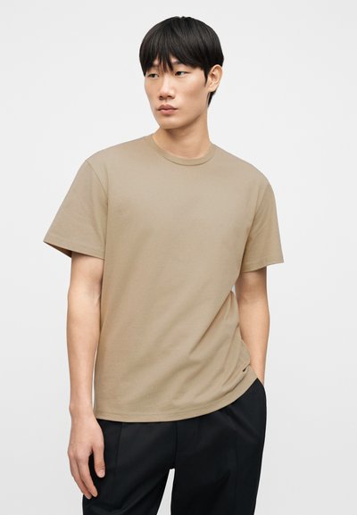 Μπεζ βαμβακερό T-shirt με κλασικό στρογγυλό λαιμόκοψη, κοντά μανίκια και άνετη γραμμή, συνδυασμένο με μαύρο παντελόνι.