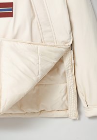 Lys beige jakke med en quiltet inderfor, med en lynlåslomme og en vandret række af dekorative striber på brystområdet.