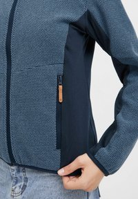 Veste pour femmes bleu marine et bleu clair avec un motif en nid d'abeille texturé, poche zippée et poignets élastiques pour un maintien sûr.