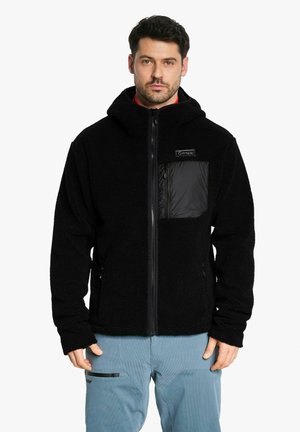 Mann trägt schwarze Fleecejacke mit Reißverschluss und Kapuze sowie hellblaue Hose und steht vor weißem Hintergrund.