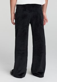 Czarne sztruksowe spodnie o szerokich nogawkach z miękką, teksturowaną powierzchnią i przeszytymi prążkami, pokazane z tyłu, z pasem i delikatnym wykończeniem u dołu.