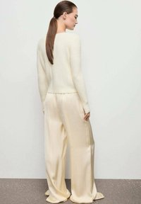 Donna con una lunga coda di cavallo castana, indossa un maglione lavorato a maglia color crema e pantaloni beige in satin a gamba larga, in piedi di profilo contro un muro chiaro e semplice.