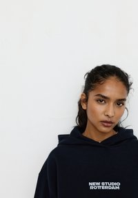 Młoda kobieta o ciemnych włosach związanych do tyłu, ubrana w granatową bluzę z kapturem z napisem „NEW STUDIO ROTTERDAM”, stoi na tle gładkiej białej ściany.