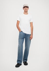 Biela tričko s krátkym rukávom, modré džíny s voľným fitom a svetlošedá čiapka s logom. Čierne topánky dotvárajú outfit.