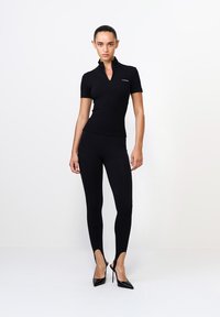 Fekete, testhez simuló póló zipzárral a gallérnál, fekete leggings-szel, amelyeknek kivágott bokái vannak, kiegészítve fekete, hegyes sarkú cipőkkel.