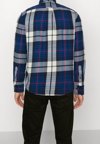 Camisa de cuadros de manga larga en tonos azules, grises y rojos. Cuenta con puños con botones, un pliegue en la parte trasera y un dobladillo curvado. El material parece suave y texturizado.