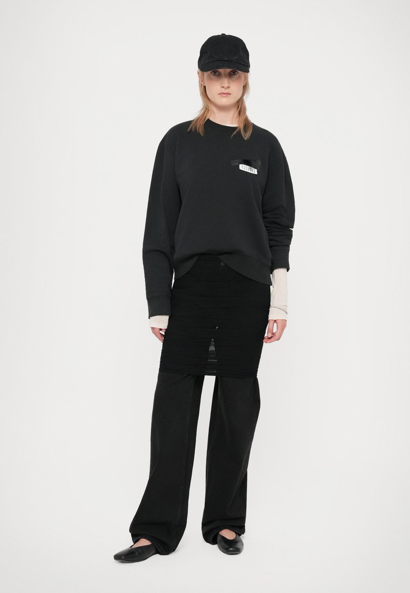 Person trägt eine schwarze Mütze, einen schwarzen Sweatshirt, einen schwarzen gerafften Rock über einer schwarzen weit geschnittenen Hose, eine beigefarbene Langarmbluse und schwarze Ballerinas vor weißem Hintergrund.