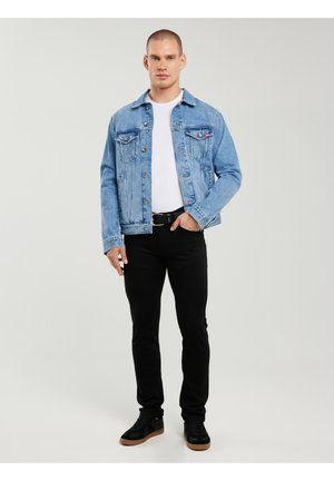 Jaunuolis stovi dėvėdamas šviesiai mėlyną džinsinę striukę, baltą marškinėlį, juodus kelnes ir juodus sportinius batelius su gumos padais, ranka kišenėje.