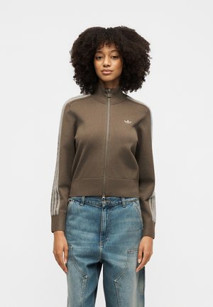 Donna con capelli ricci che indossa una giacca marrone con zip e strisce sulle maniche e jeans blu a vita alta, in piedi davanti a uno sfondo semplice.
