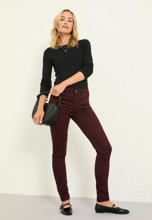 Frau in einem schwarzen langärmligen Oberteil, bordeauxfarbenen Skinny-Hosen, schwarzen Ballerinas und mit einer kleinen schwarzen Handtasche, die an einer schlichten Wand steht.