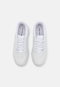 Ett par vita Lacoste-sneakers med snörning, perforerade detaljer på tån och grönt logotyp inuti, sedd uppifrån på en grå bakgrund.