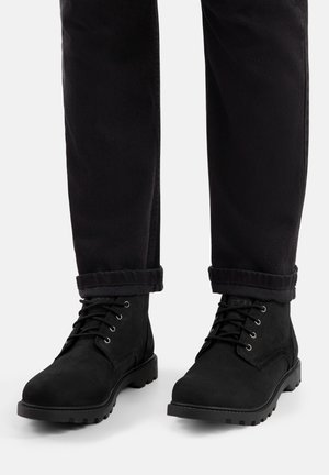 Crne gležnjače od brušene kože s teksturiranim završetkom i gumenim potplatima s profilom, uparene s crnim hlačama s podvrnutim rubovima. Detalji s rupicama i vezicama.