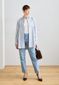 Camicia a righe blu e bianche con bottoni, camicia bianca aderente sotto, jeans azzurri chiari, tacchi a punta neri e una piccola borsa marrone.