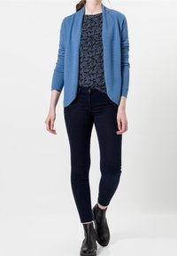 Cárdigan azul claro con un patrón texturizado, superpuesto a una blusa de estampado paisley oscuro, combinado con jeans ajustados oscuros y botas negras tipo ankle.