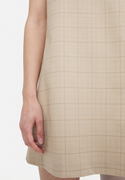 Beiges karierter Kleid mit einer schlichten Silhouette, das ein strukturiertes Muster und einen leicht ausgestellten Saum aufweist. Eine Hand liegt auf dem Stoff.