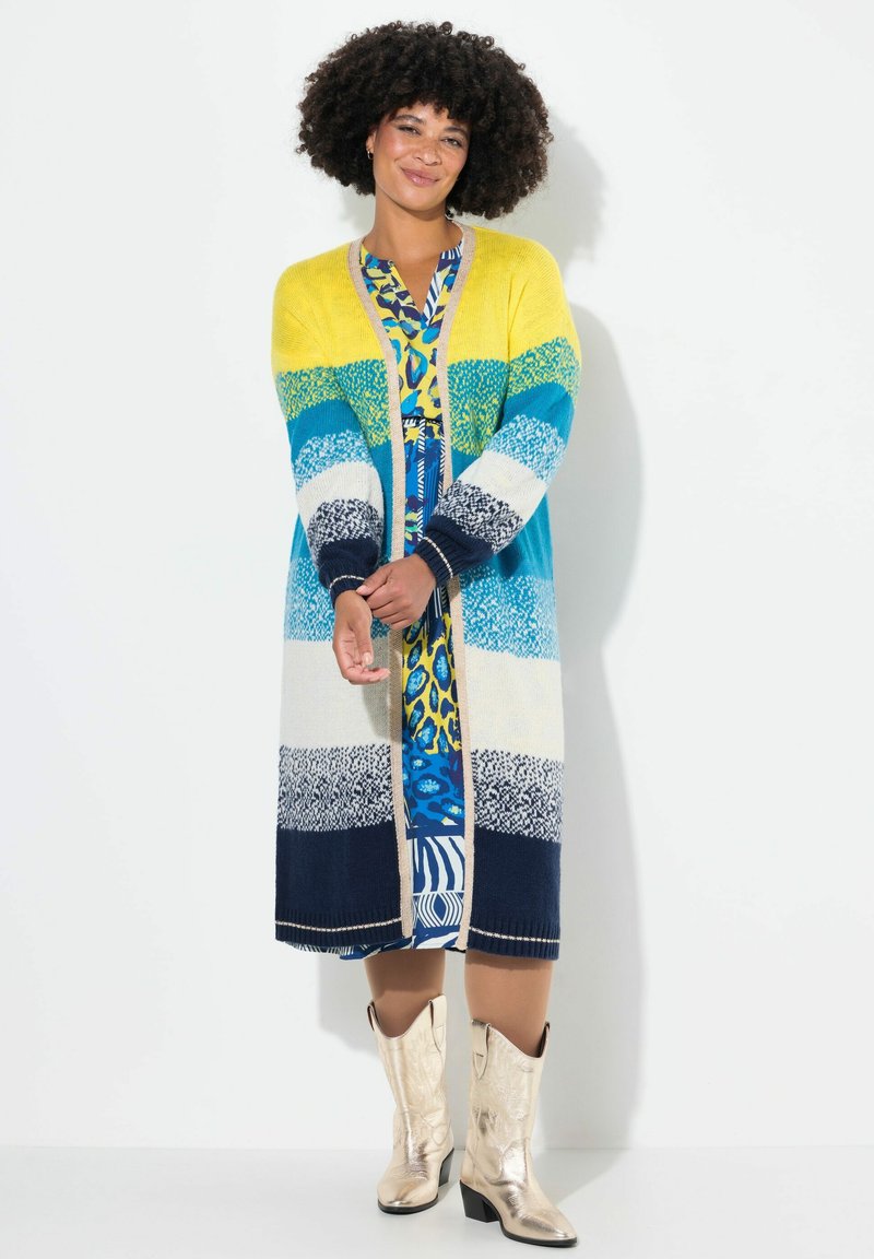 Stricklongcardigan mit gelben, türkisfarbenen, weißen und marineblauen Streifen, mit gerippten Bündchen. Über ein gemustertes, blaues Kleid und goldene Stiefel getragen.